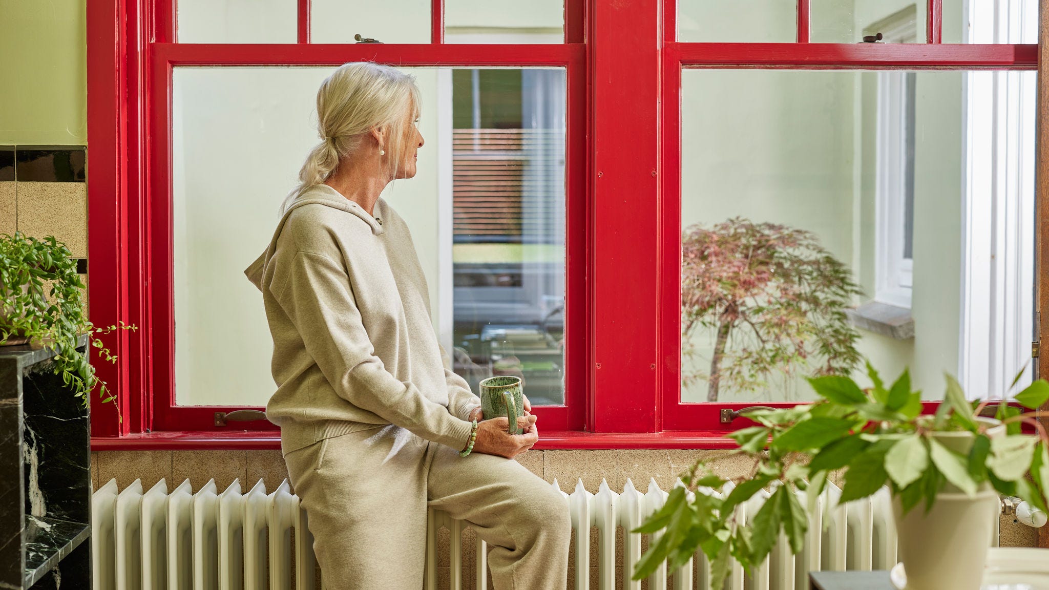 Femme assise sur un radiateur près d’une grande fenêtre, profitant d’une habitation chaleureuse et éco-énergétique