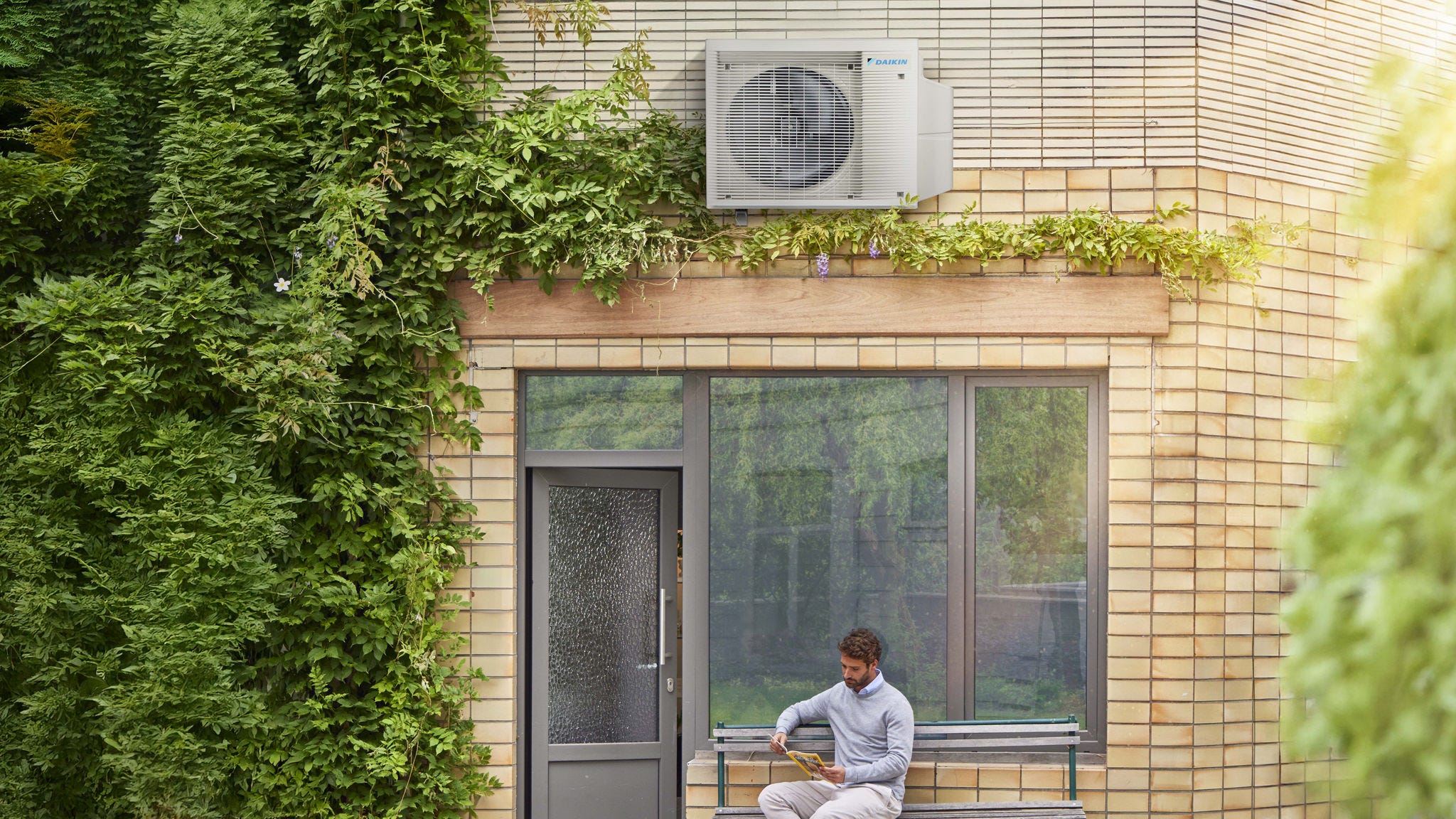 Homme lisant sur un banc à l'extérieur de sa maison, sous une pompe à chaleur aérothermique Daikin.