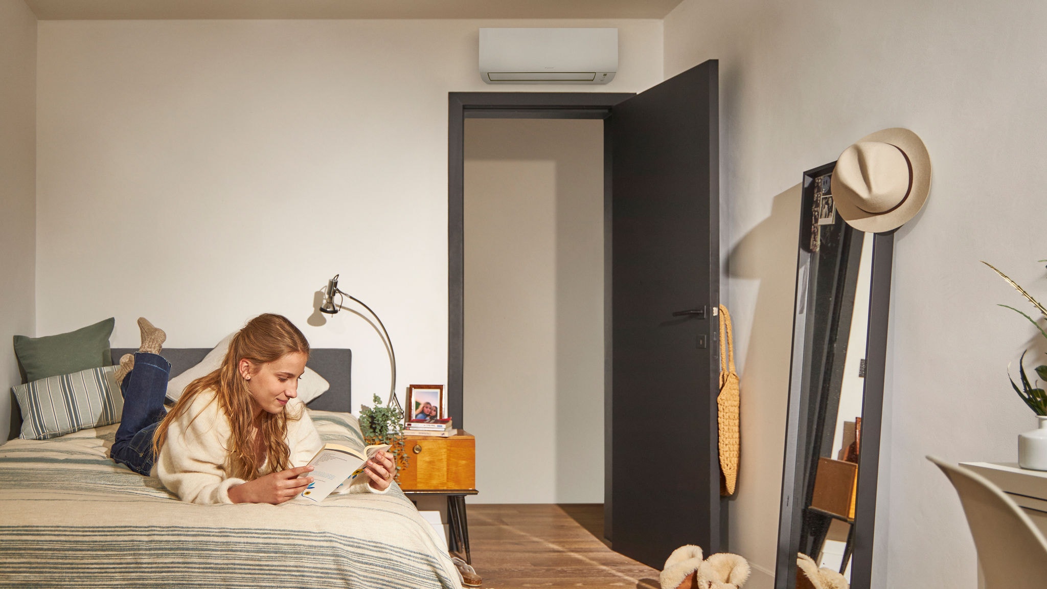 Femme lisant sur un lit dans une chambre confortable, avec une unité de climatisation de type pompe à chaleur installée au-dessus de la porte et assurant un conditionnement de l'air éco-énergétique