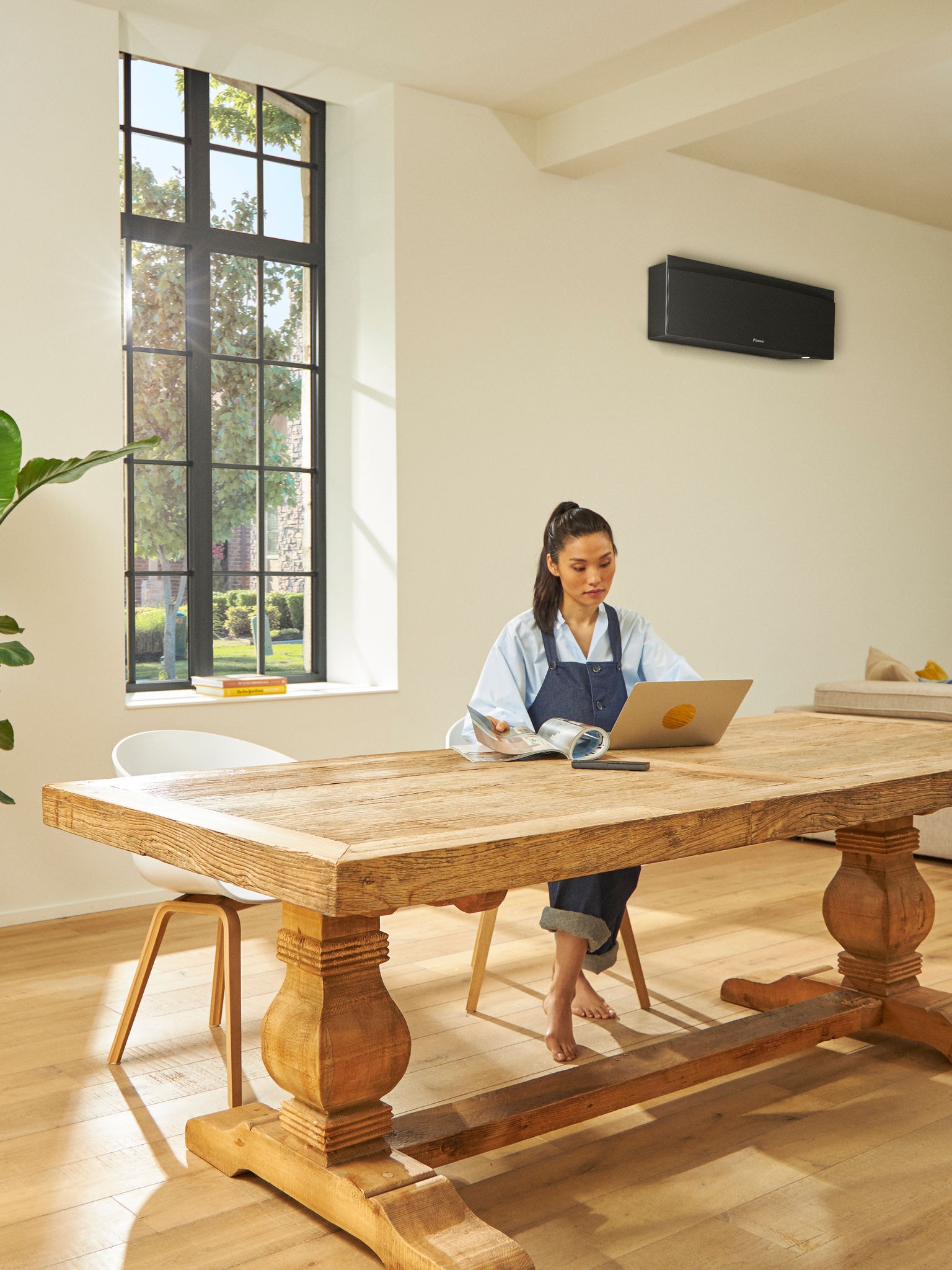 Femme assise à une table avec un ordinateur portable, le soleil traversant une fenêtre au cadre noir, et une élégante pompe à chaleur murale noire discrète en arrière‑plan