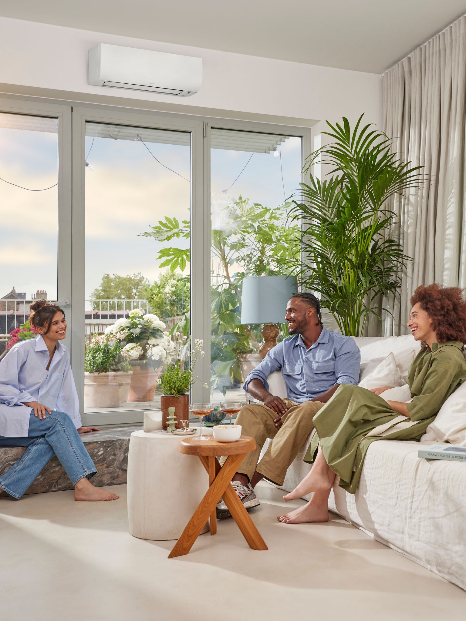 Un homme et deux femmes discutant par une journée ensoleillée dans une pièce lumineuse aux murs blancs, équipée d’une pompe à chaleur murale blanche discrète installée au‑dessus d’une grande baie vitrée
