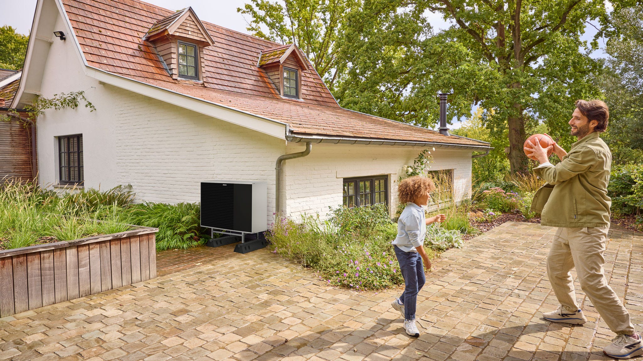 Père jouant au ballon avec son enfant dans un jardin, avec une pompe à chaleur installée à proximité