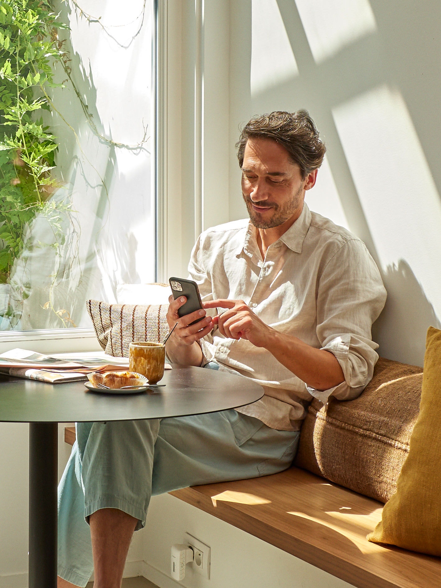 Homme se détendant et regardant son téléphone, assis à table devant une tasse de café et de la nourriture.