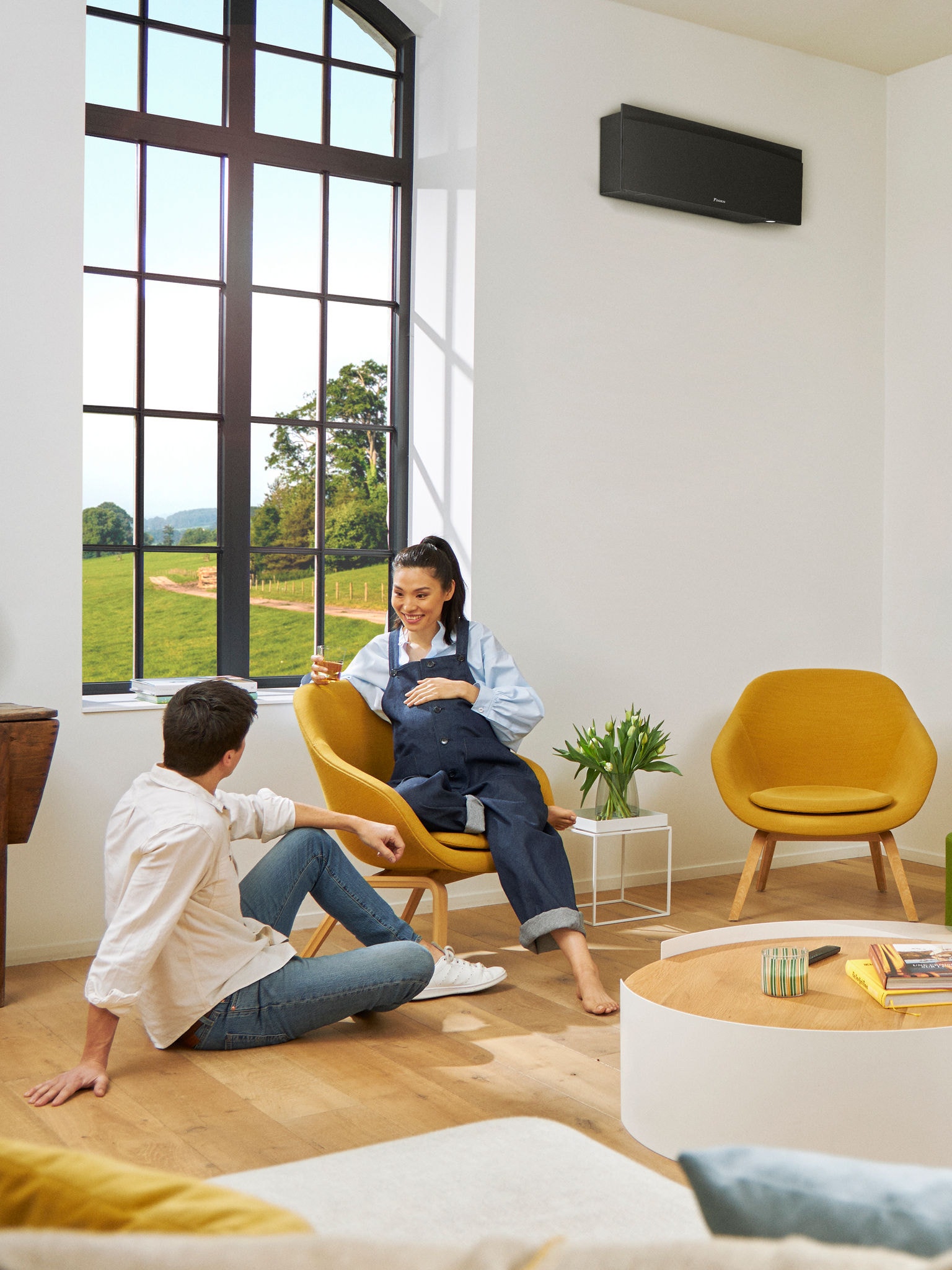 Un homme et une femme assis devant une grande fenêtre dans une pièce lumineuse, avec une pompe à chaleur murale noire installée sur le mur derrière eux