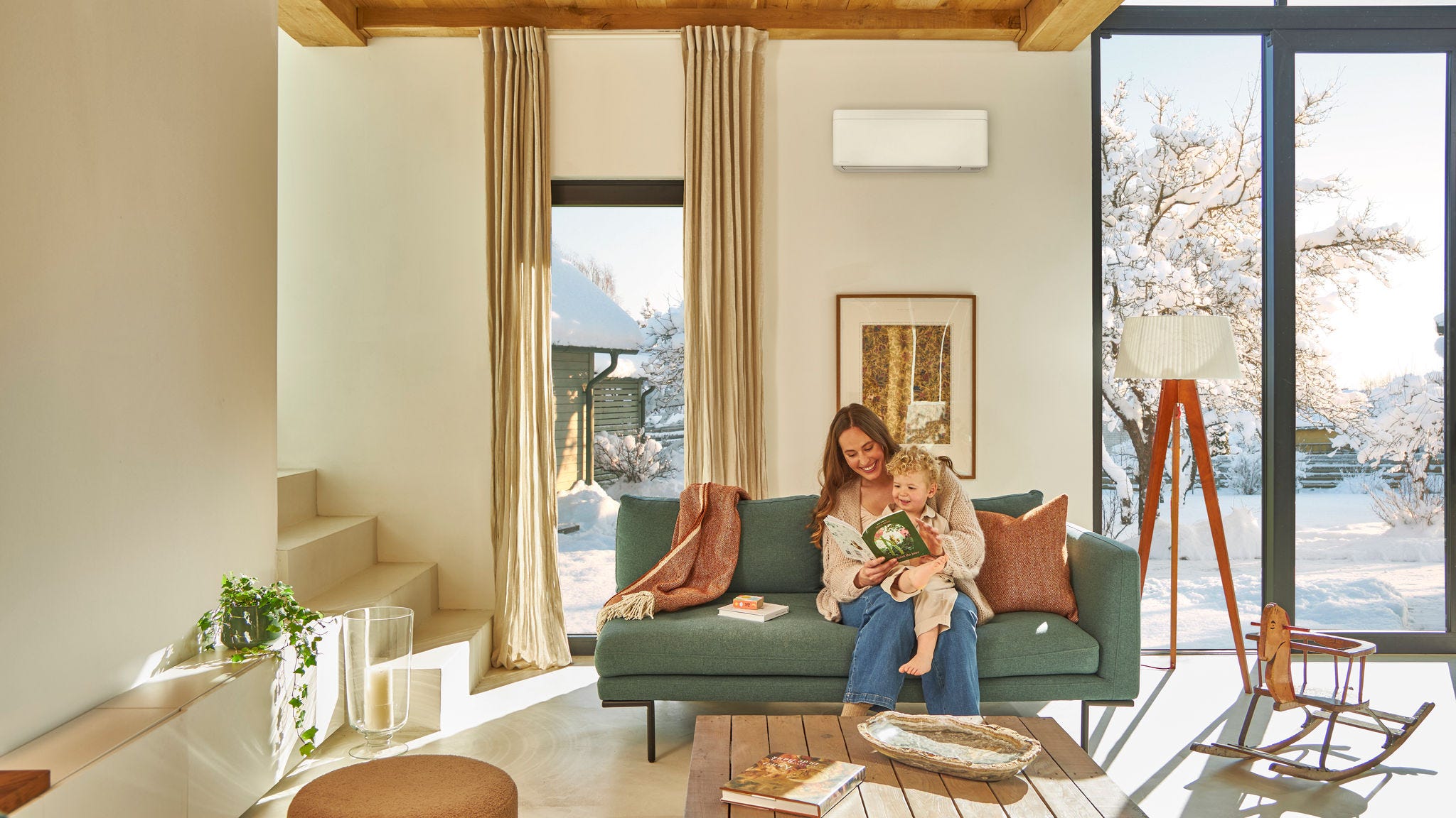 Mère assise sur le canapé sous une pompe à chaleur air-air murale, lisant un livre à son enfant assis sur ses genoux, profitant de la chaleur alors qu'il neige dehors.
